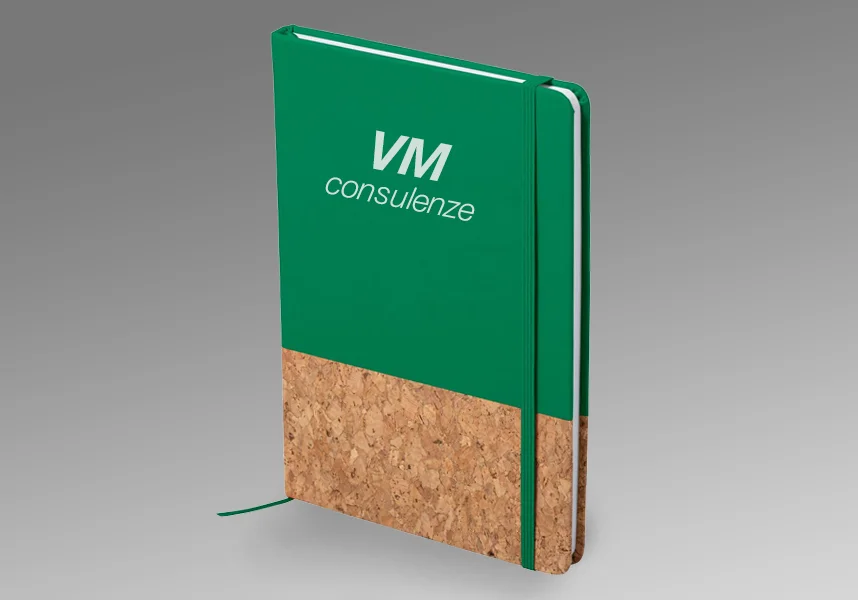 Un quaderno con copertina rigida verde e sughero con VM consulenze stampate sul fronte, dotato di elastico e segnalibro a nastro, perfetto come gadget promozionale, esposto su sfondo grigio.