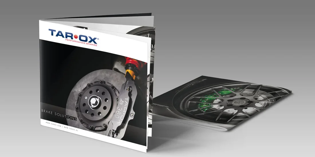 Sono esposte due brochure automobilistiche: una è aperta e mostra l'immagine di un disco dei freni sulla copertina, mentre l'altra è distesa e rivela parte della ruota di un'auto e del sistema frenante al suo interno.