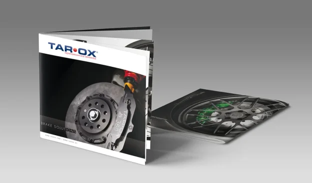 Sono esposte due brochure automobilistiche: una è aperta e mostra l'immagine di un disco dei freni sulla copertina, mentre l'altra è distesa e rivela parte della ruota di un'auto e del sistema frenante al suo interno.