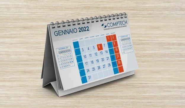 Un calendario da tavolo aperto fino a gennaio 2022, con date evidenziate in blu e rosso, marchiato con il logo Comptech, posto su una superficie di legno.