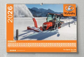 Un trattore Kubota rimuove la neve da una strada rurale con un accessorio spazzaneve; sullo sfondo sono visibili montagne e campi. Bold Tipografia mette in evidenza l'immagine su un calendario da parete 2026 per il mese di gennaio.