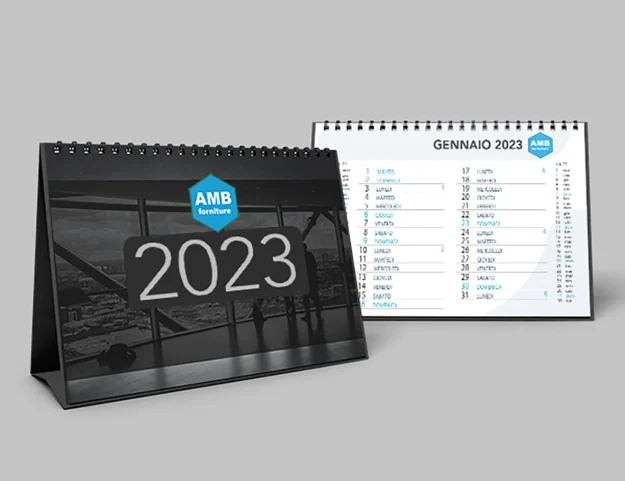 calendario_stampa_2023 Sono esposti due calendari da tavolo per il 2023, uno che mostra l'anno con il logo di AMB Forniture, l'altro aperto a gennaio con date e nomi elencati in italiano.