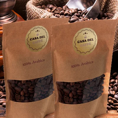 Due sacchetti marroni con l'etichetta CASA DEL CAFFÈ 100% Arabica sono esposti davanti a un mucchio di chicchi di caffè e a un misurino di metallo.