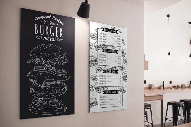 Due lavagne da menu montate a parete in un ristorante; una con l'illustrazione di un hamburger e il testo Burger Menu, l'altra che elenca le opzioni e i prezzi degli hamburger. Sono visibili sgabelli e tavoli in legno.