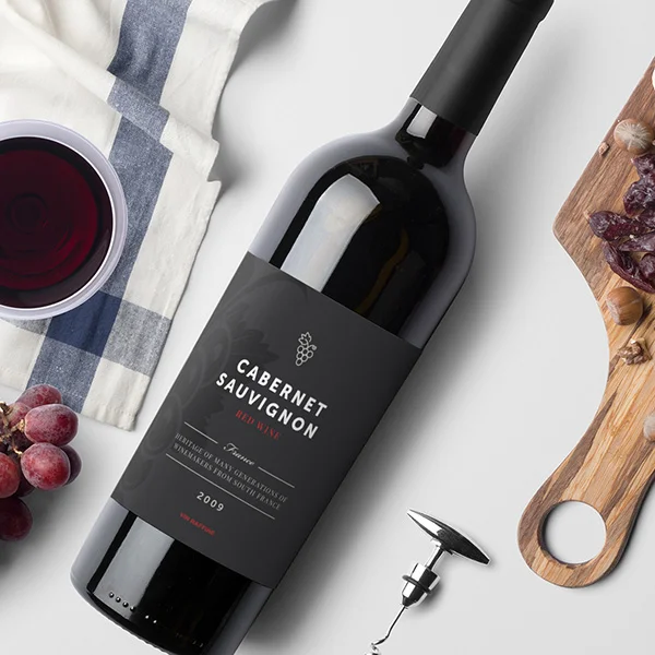 Una bottiglia di Cabernet Sauvignon su un tavolo con un bicchiere di vino rosso, uva, un cavatappi e una tavola di legno con affettati.