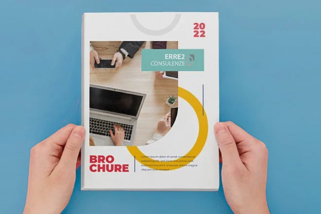Una persona tiene in mano un opuscolo del 2022 intitolato BROCHURE con in copertina l'immagine di persone che lavorano a una scrivania con computer portatili e documenti.